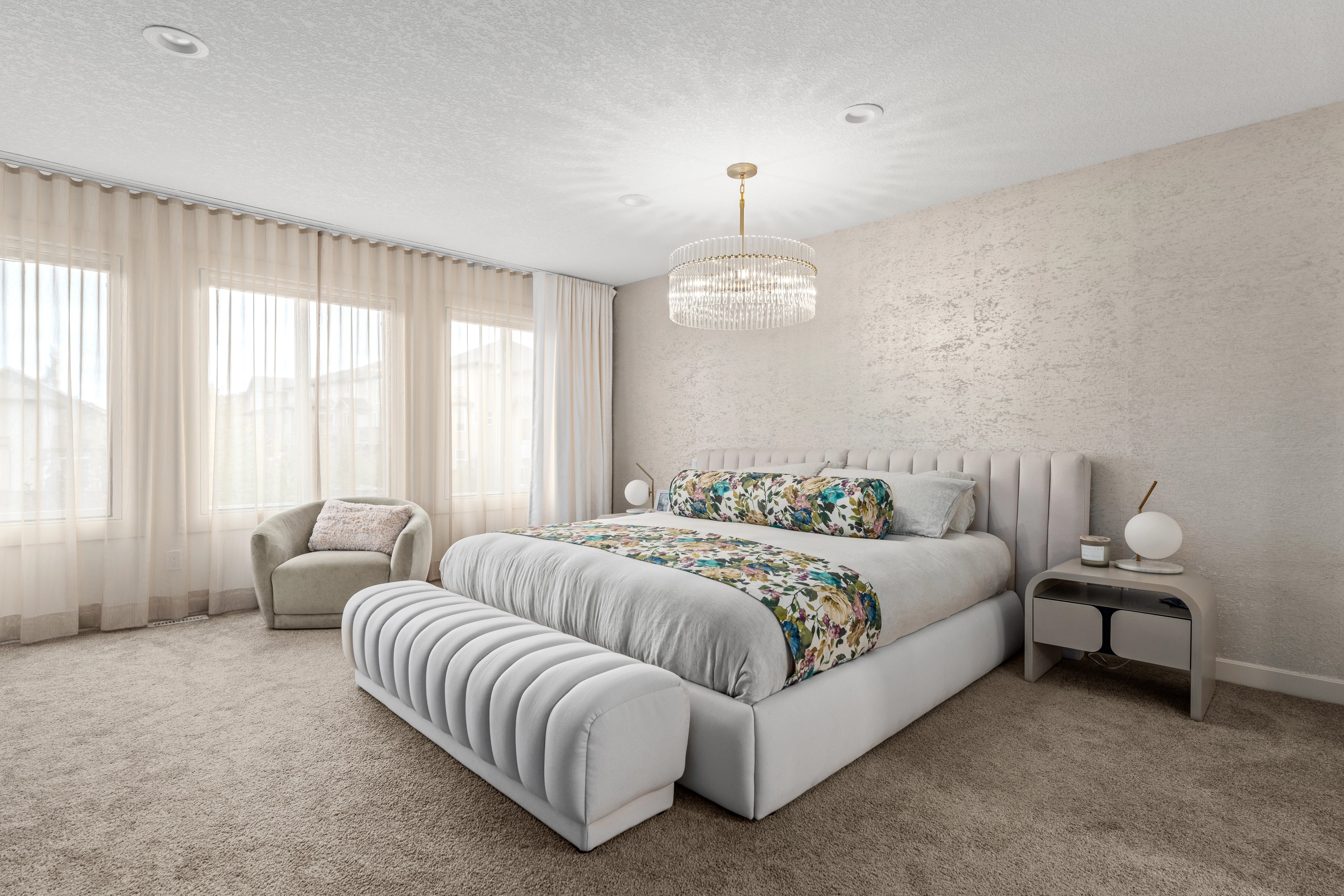 Wide view of the primary bedroom with a crystal chandelier, floral bedding, and a channel-tufted bench