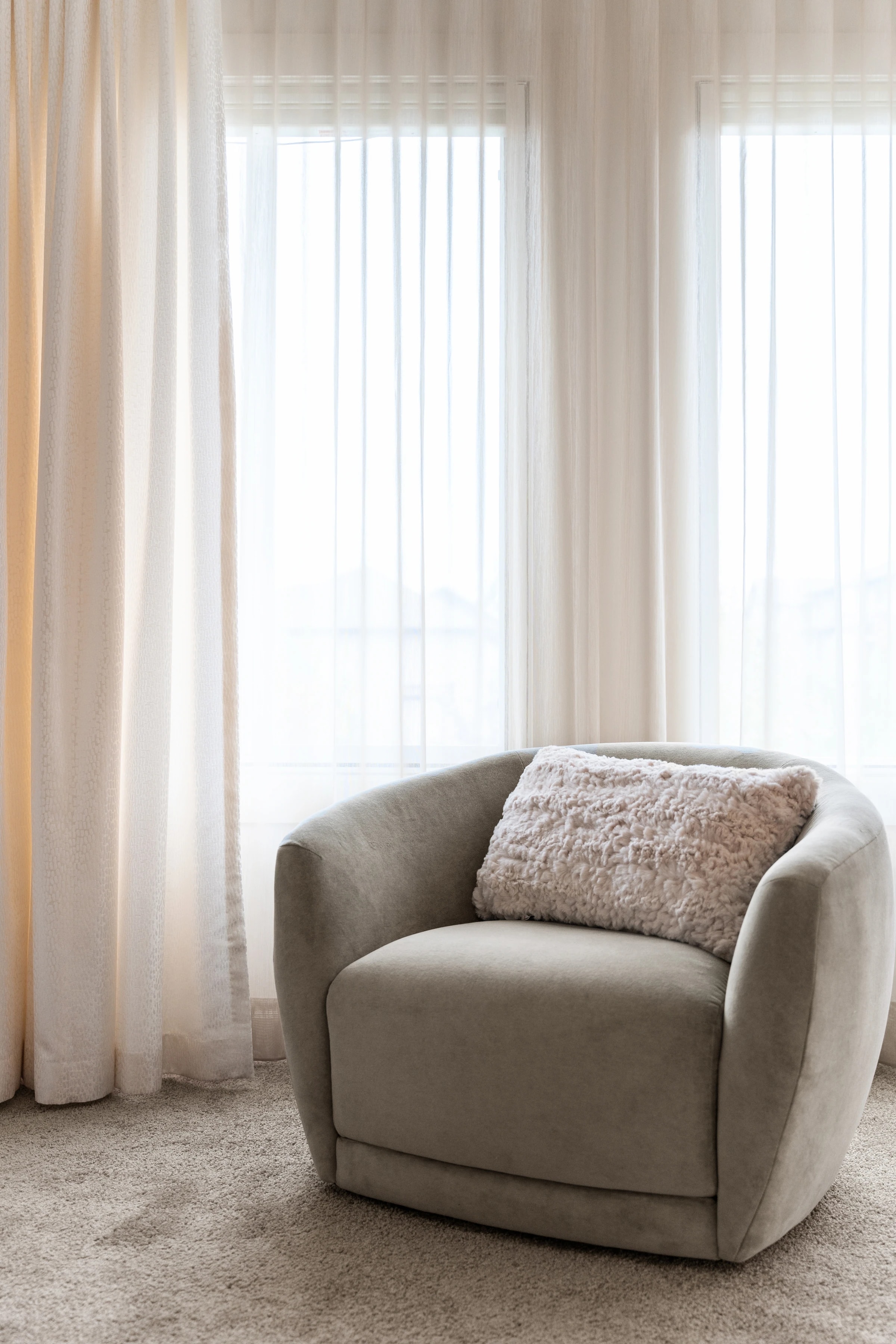 Cream swivel armchair beside sheer-curtained windows in the primary suite