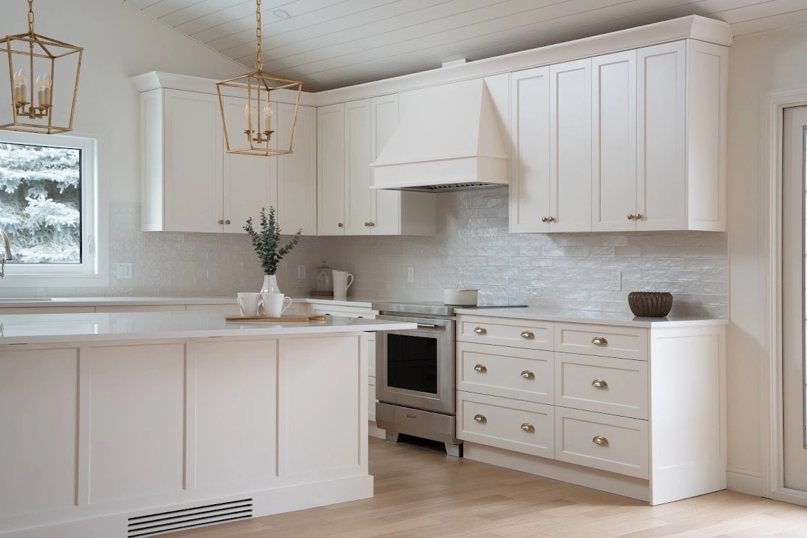 White shaker kitchen with brass lantern pendants, a painted range hood, and a window onto snowy spruce trees