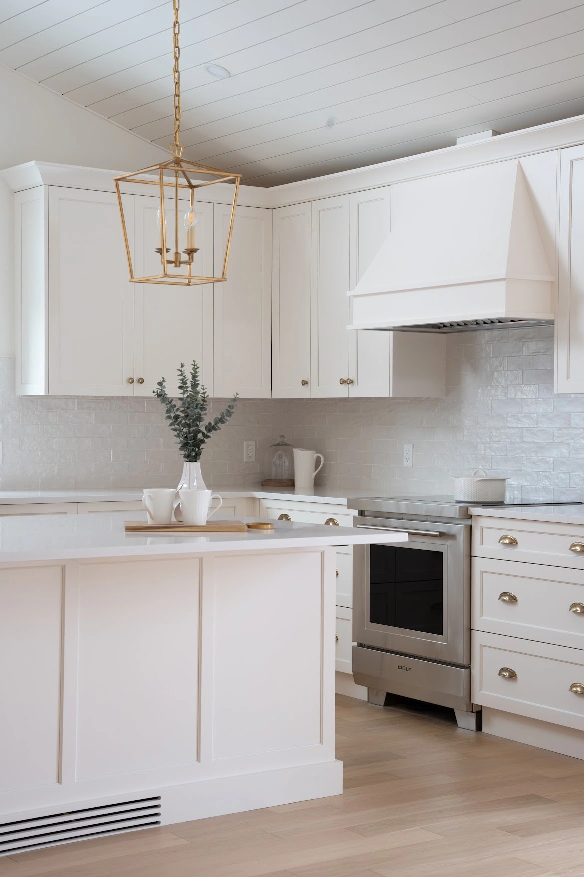 Brass lantern pendant above a white island styled with eucalyptus and mugs, with the range and tile backsplash beyond