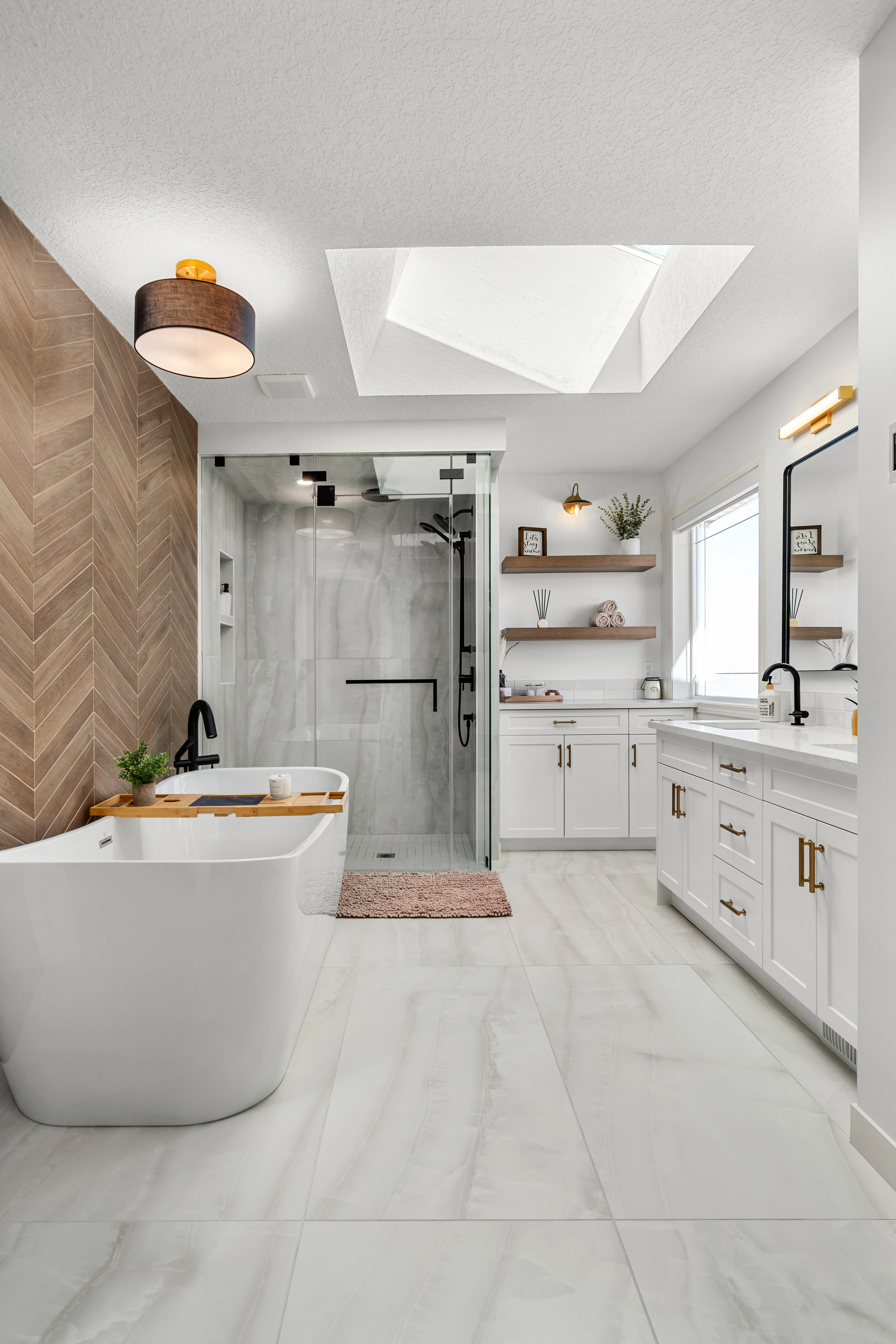 Wide view of a skylit primary ensuite with a freestanding tub, glass shower, and dual vanity
