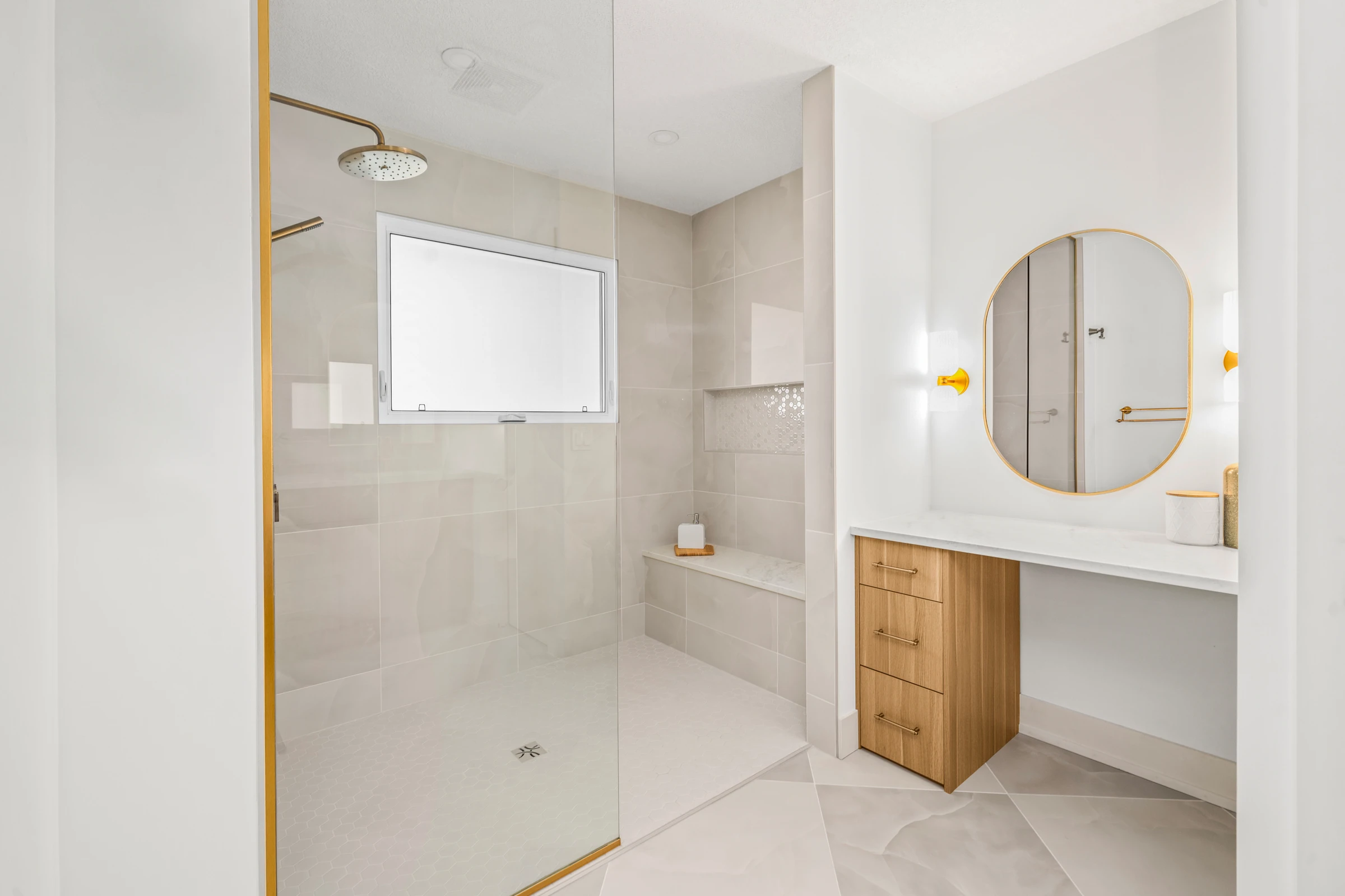 Glass-enclosed shower beside a small vanity nook with a round gold mirror