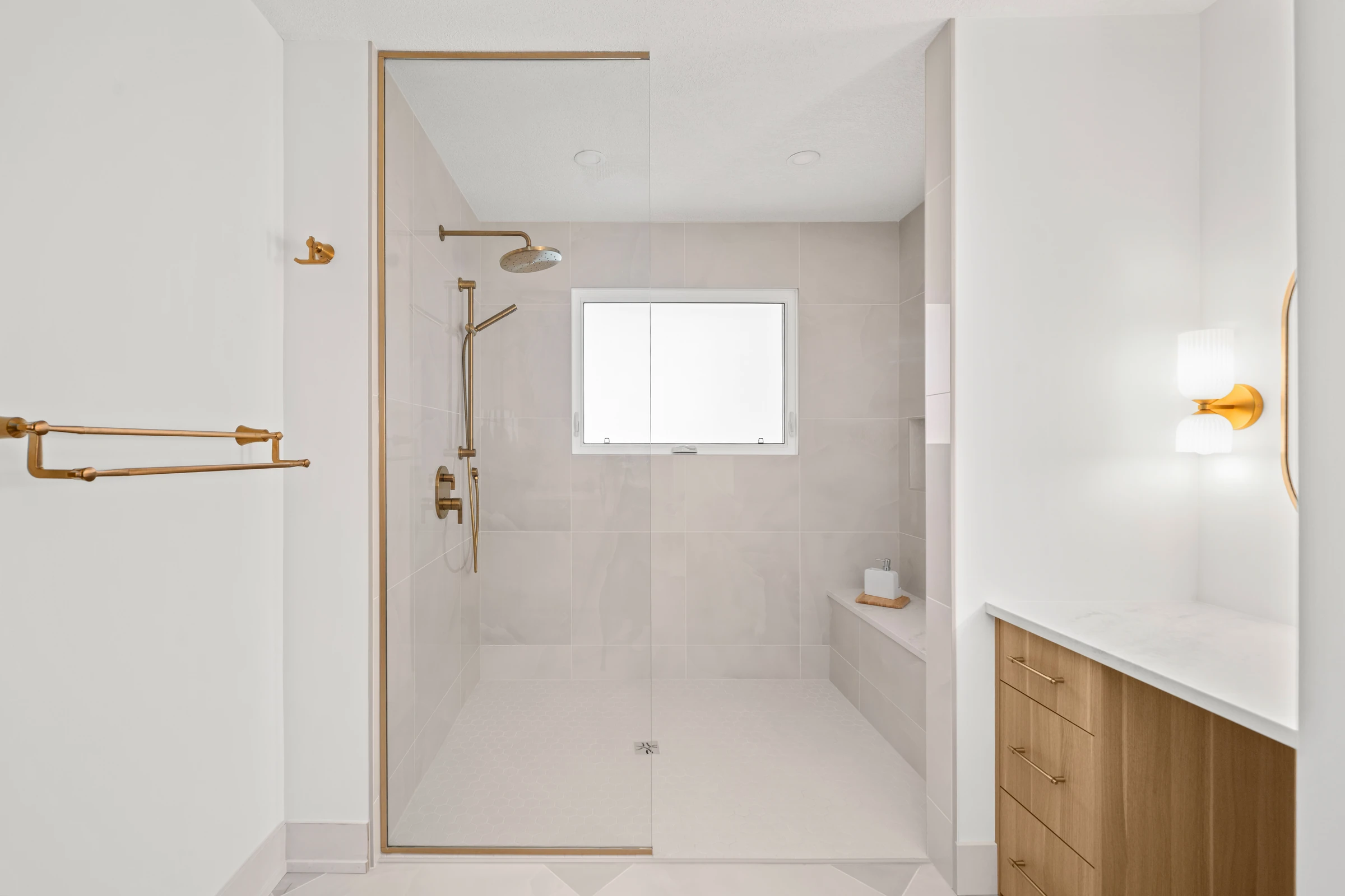 Glass shower with gold hardware beside a wood vanity in a bright white ensuite
