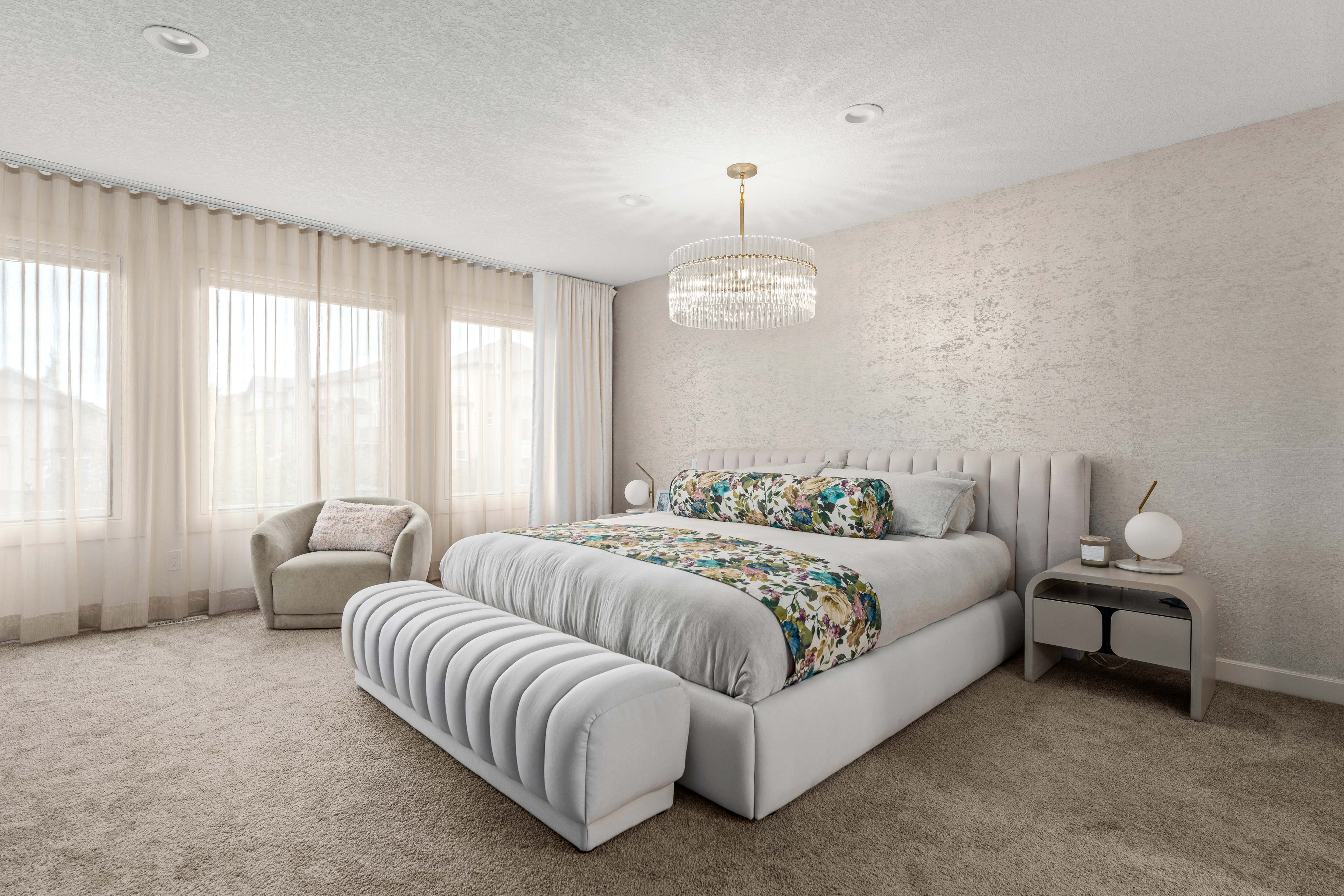 Wide view of the primary bedroom with a crystal chandelier, floral bedding, and a channel-tufted bench